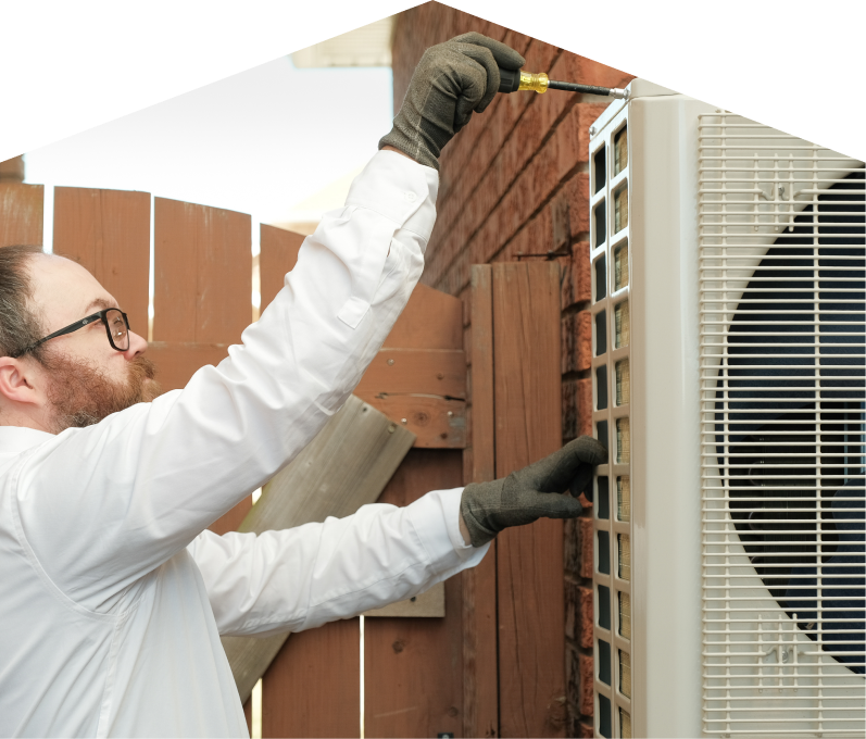 Mersey service personnel working on an AC condenser, using a screwdriver to secure the unit's panel outdoors beside a wooden fence.