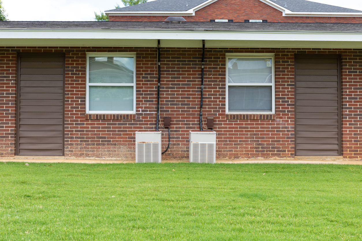 Exterior view of a generic apartment building with older model HVAC heat pumps, twin windows, green grassy lawn, creative copy space Exterior view of a generic apartment building with older model HVAC heat pumps, twin windows, green grassy lawn, creative copy space